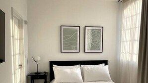 A bedroom with a bed, two framed abstract paintings, a nightstand with a lamp, and a window with curtains.