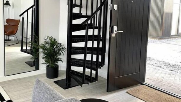 A modern interior with a black spiral staircase, a dark wooden door, and a large mirror reflecting a room with a chair and a plant.