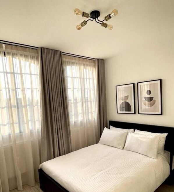 A hotel room with a bed, curtains, a ceiling light fixture, and two framed pictures on the wall. - OneFiveSix Unique ApartHotel 2 Bed (Birmingham)