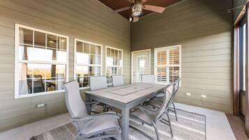 A covered patio with a table and chairs, large windows with blinds, and a ceiling fan.