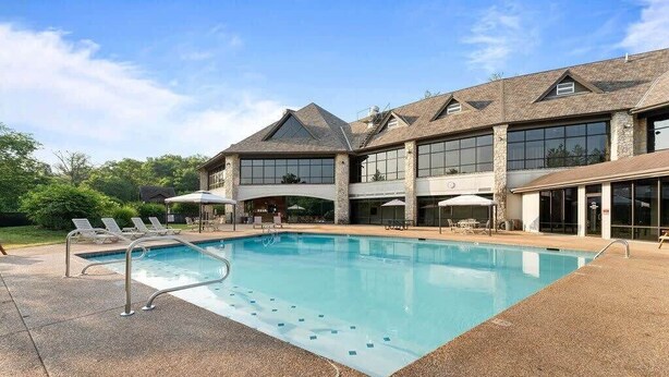 Une piscine avec des chaises longues, un patio couvert et un grand bâtiment doté de nombreuses fenêtres et d’une façade en pierre.