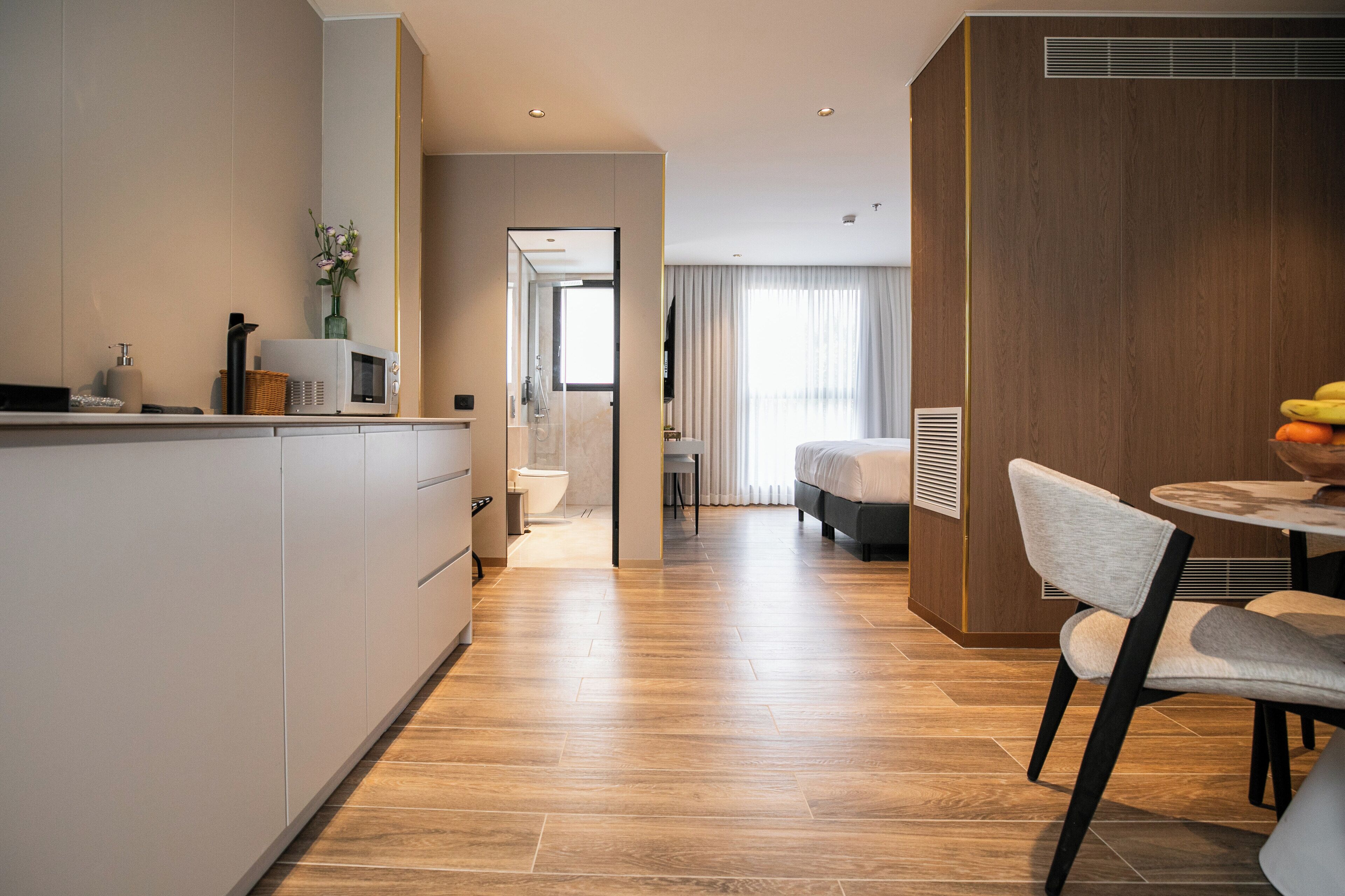 A modern hotel room with a wooden floor, a dining area with a round table and chairs, a large bed, and a bathroom visible through an open door.