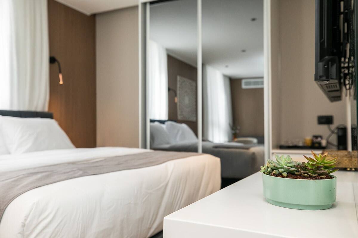 A modern hotel room with a bed, a small table with a potted plant, and a large mirror reflecting the room.