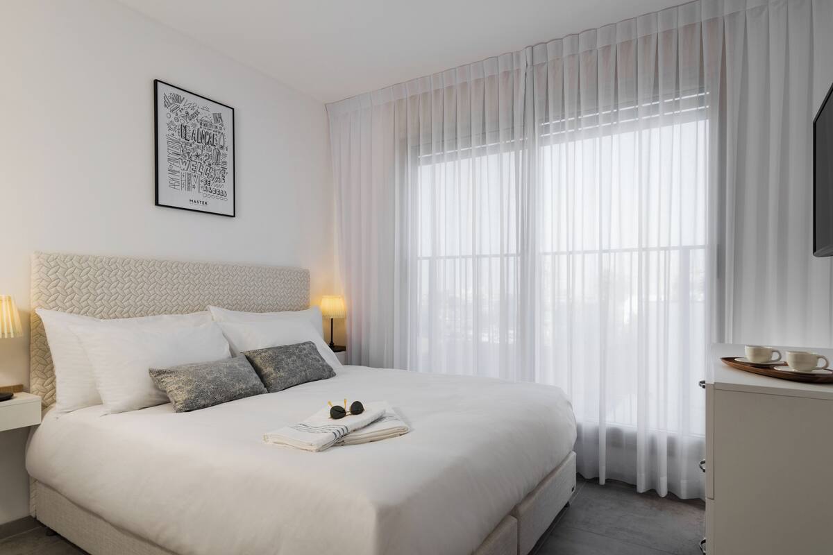 A neatly made bed with pillows, a bedside table with a lamp, a framed picture on the wall, and a window with sheer curtains.