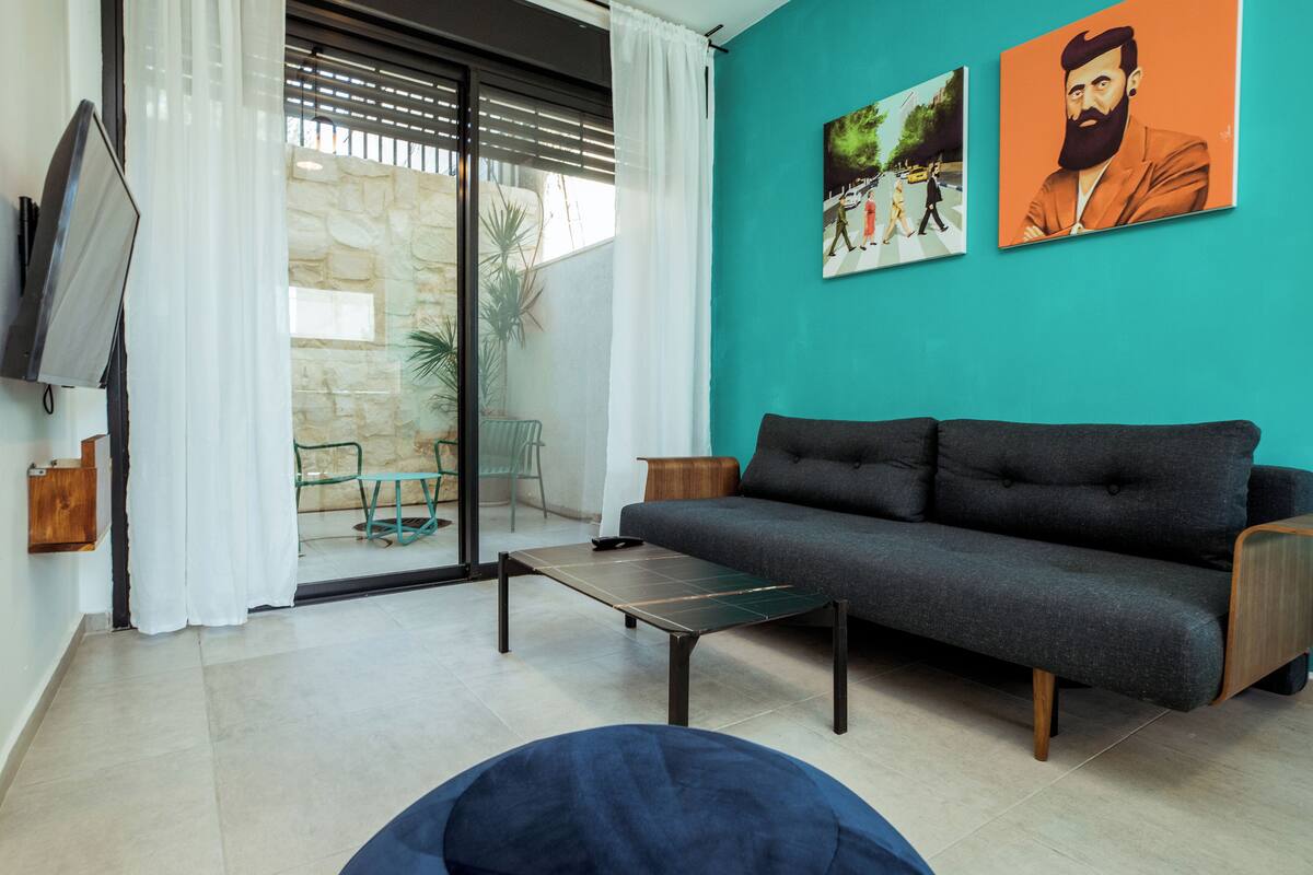 A living room with a sofa, a coffee table, and a TV. There are two paintings on the wall and a window with curtains.