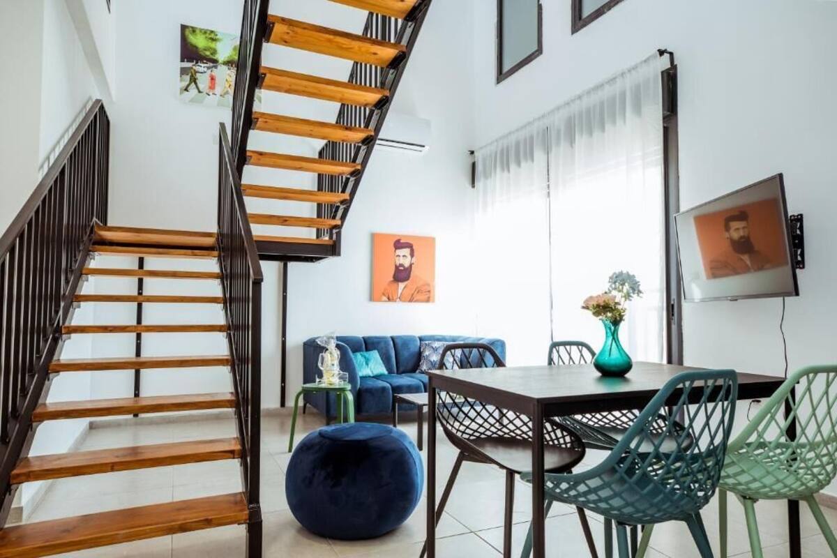 A staircase with wooden steps and black railings, a blue sofa with patterned cushions, a dining table with chairs, and a vase with flowers.
