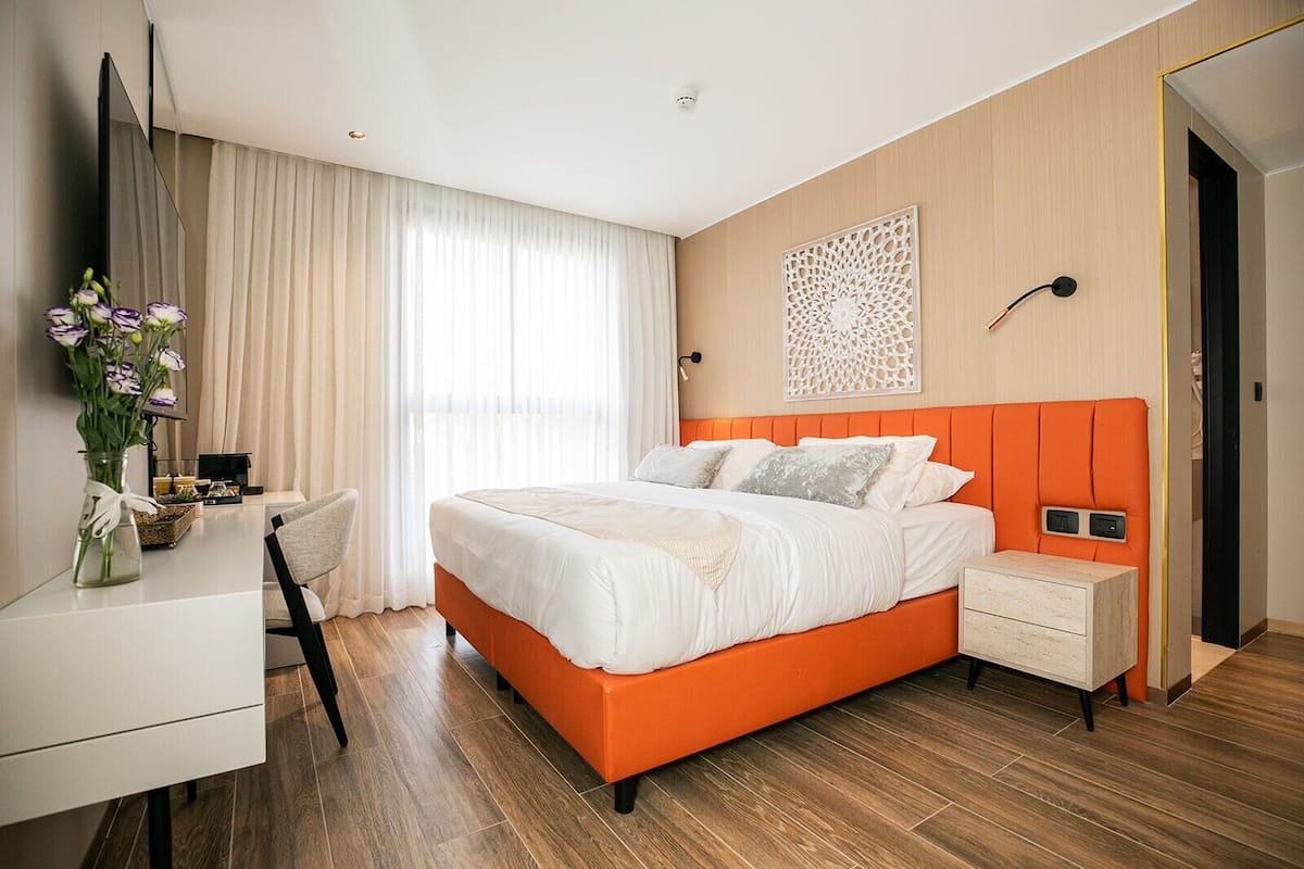 A modern hotel room with a large bed, a bedside table, a desk with a chair, and a decorative wall piece.