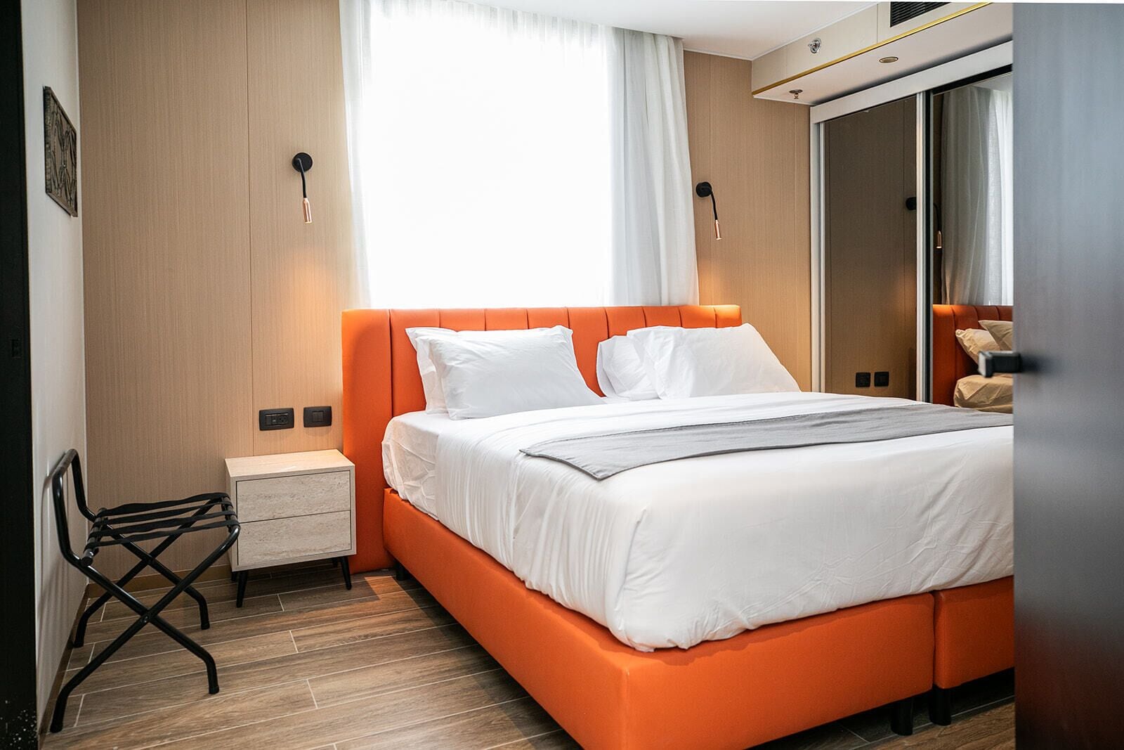 A modern hotel room with a large bed, a nightstand, a chair, and a wardrobe.
