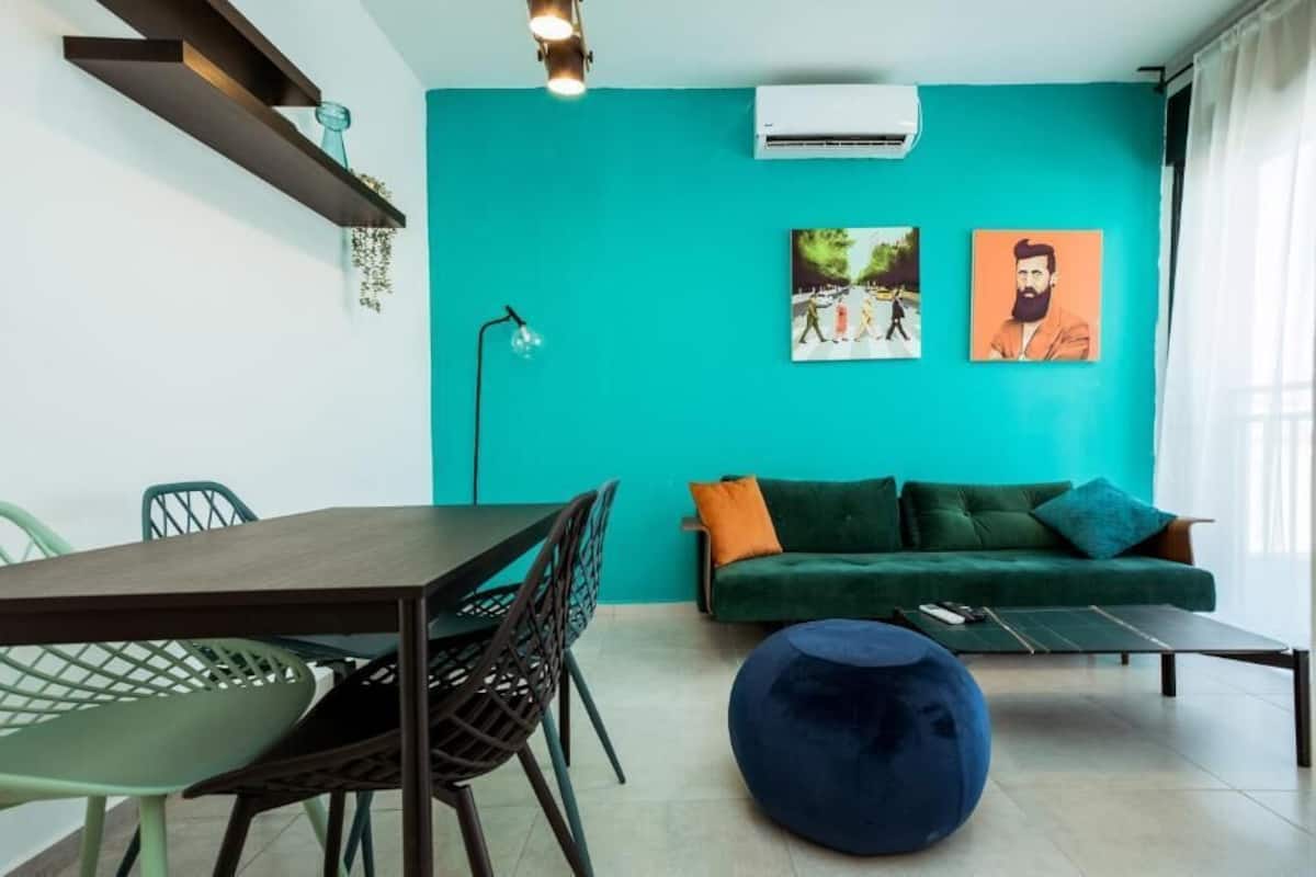 A modern living space with a teal accent wall, a green sofa with orange cushions, a dining table with chairs, and a blue ottoman.