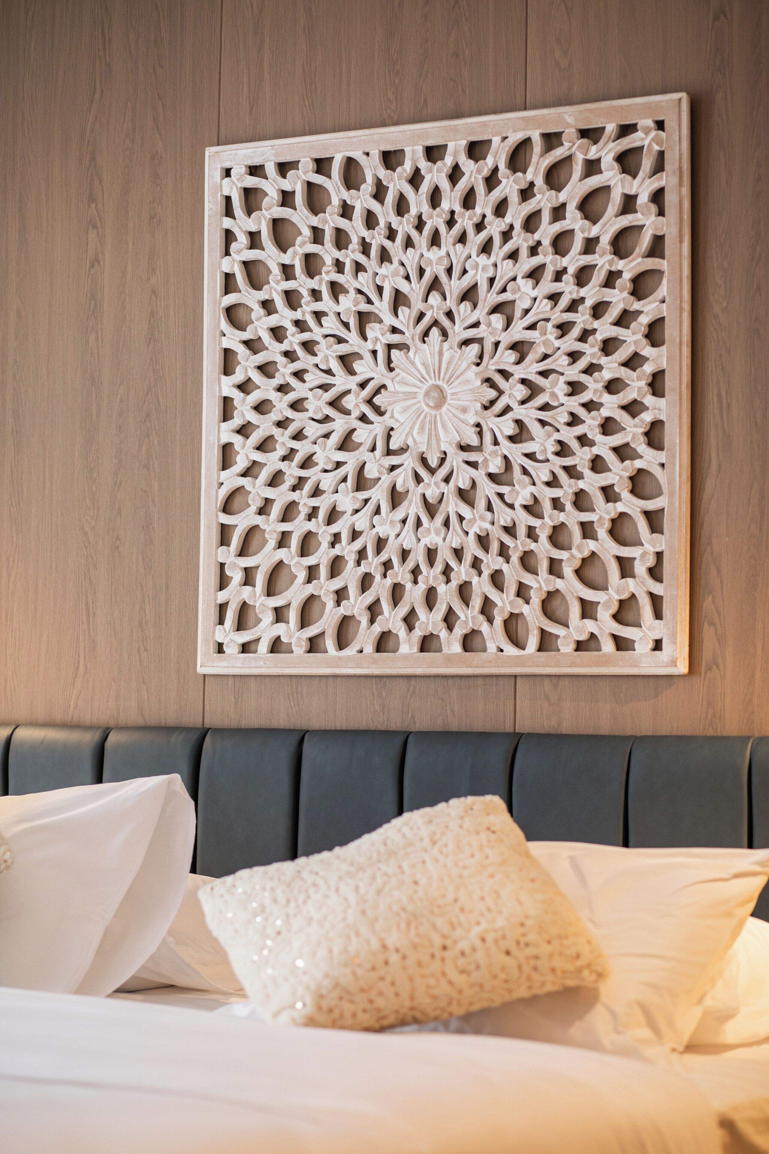 A wall-mounted decorative panel with intricate patterns, a bed with grey upholstered headboard, and white bedding with decorative pillows.