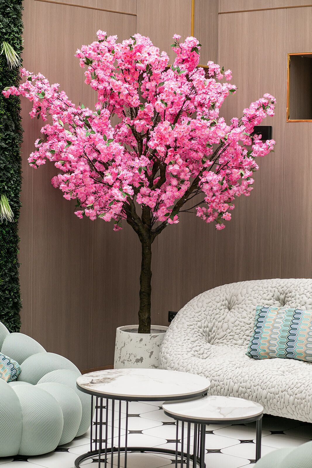 A room with a large artificial pink tree, a white sofa with patterned cushions, and a round table with a marble top.