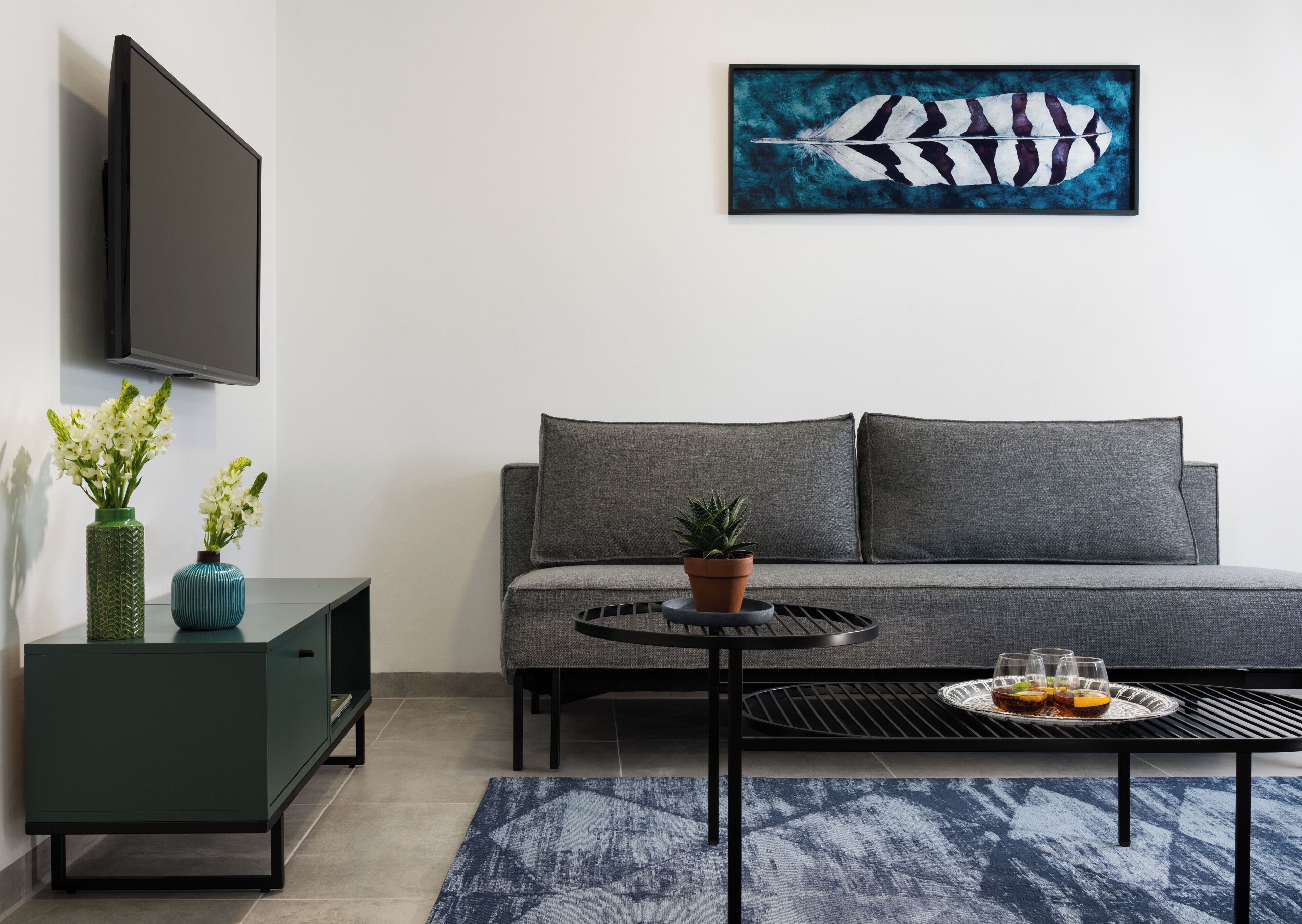 A modern living room with a grey sofa, a black coffee table with a tray of drinks, a green and blue vase with flowers, and a large painting of a feather on the wall.