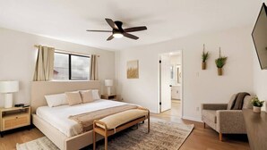 A modern bedroom with a large bed, bedside tables, a ceiling fan, a window with curtains, a door leading to another room, and a small seating area with a chair and a plant. - Radiant & Refined | Stylish 4BR Home in Round Rock (Round Rock)