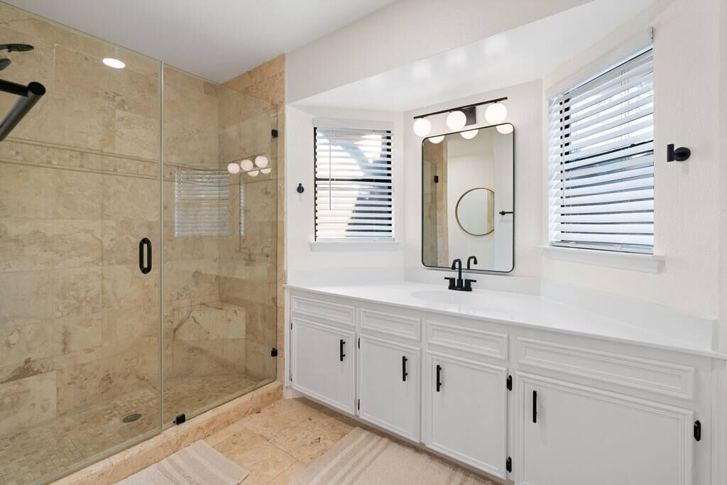 Une salle de bain moderne dotée d’une douche à parois de verre, d’un meuble-lavabo avec miroir et éclairage, et d’une fenêtre avec stores.