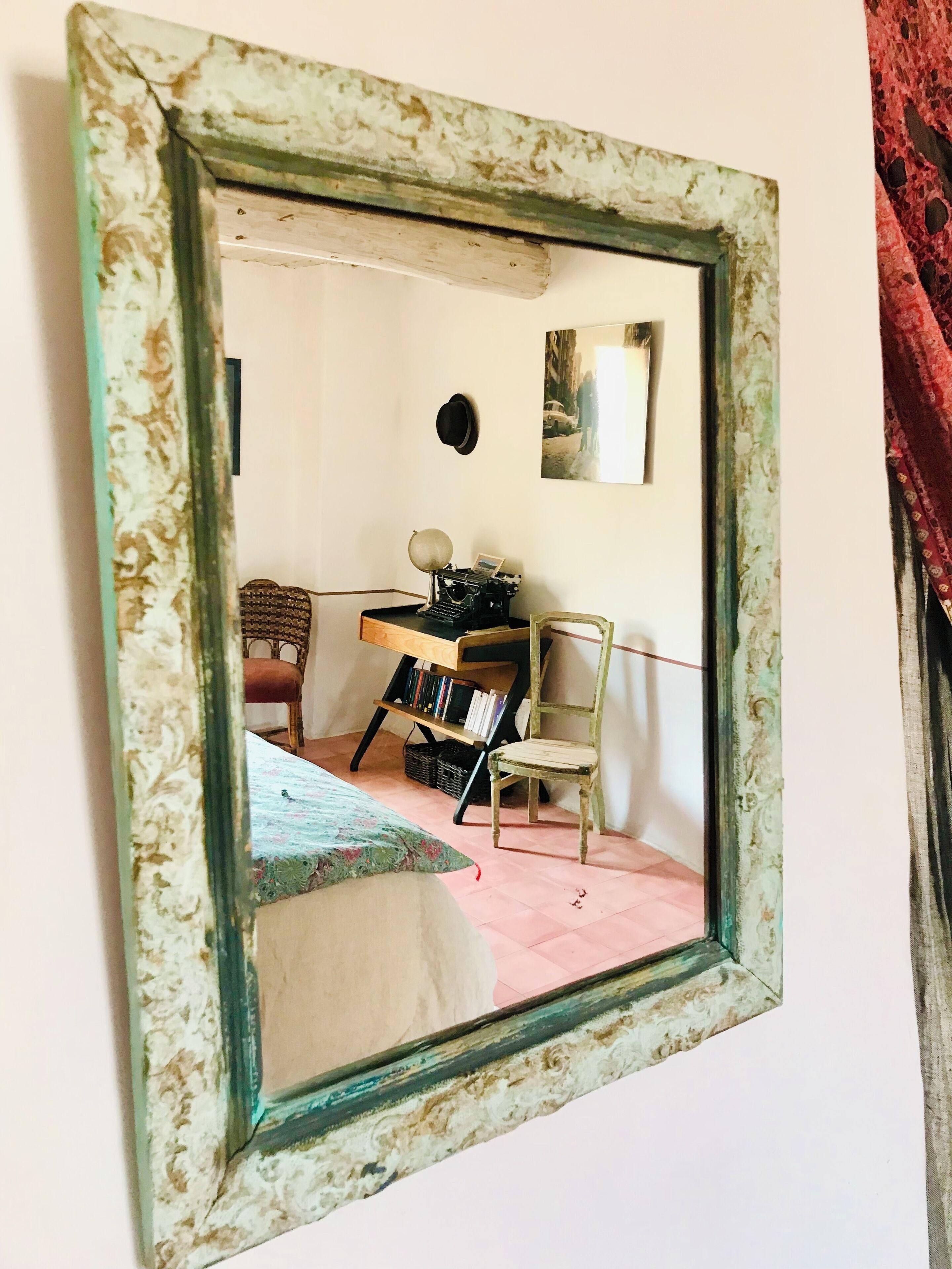 Une pièce avec un miroir ancien qui reflète un bureau, une chaise, une bibliothèque et un lit recouvert d’une couvre-lit à motifs floraux.
