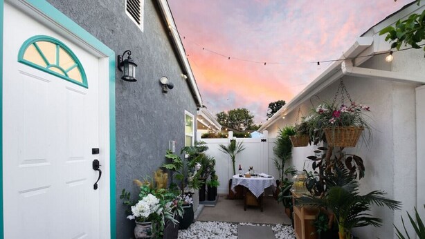 A narrow outdoor patio with a white door, potted plants, a table with chairs, and a sunset view.