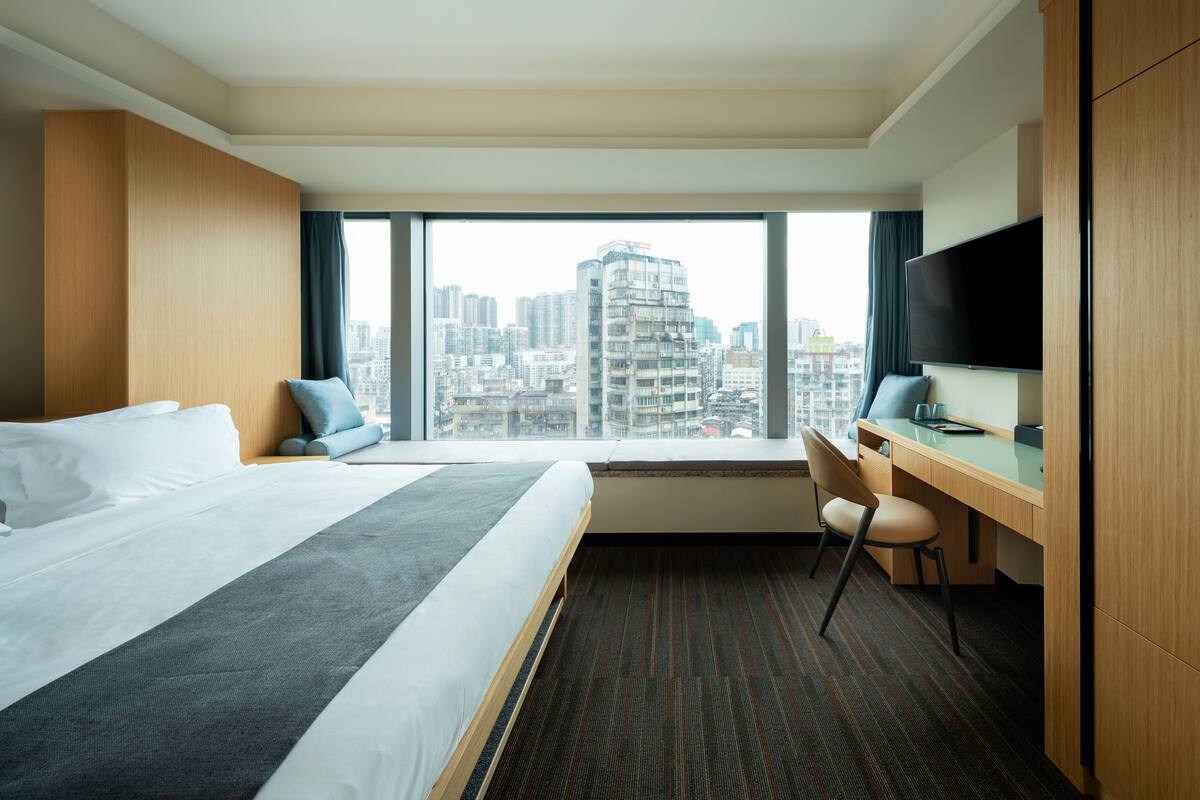 A hotel room with a large bed, a desk, a chair, a TV, and a city view.