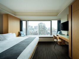Habitación de hotel con una cama grande, un escritorio, una silla, televisión y vista a la ciudad.