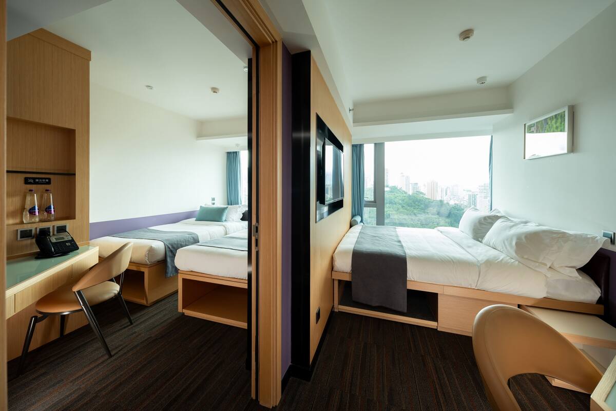 A hotel room with two beds, a desk, and a view of a cityscape.