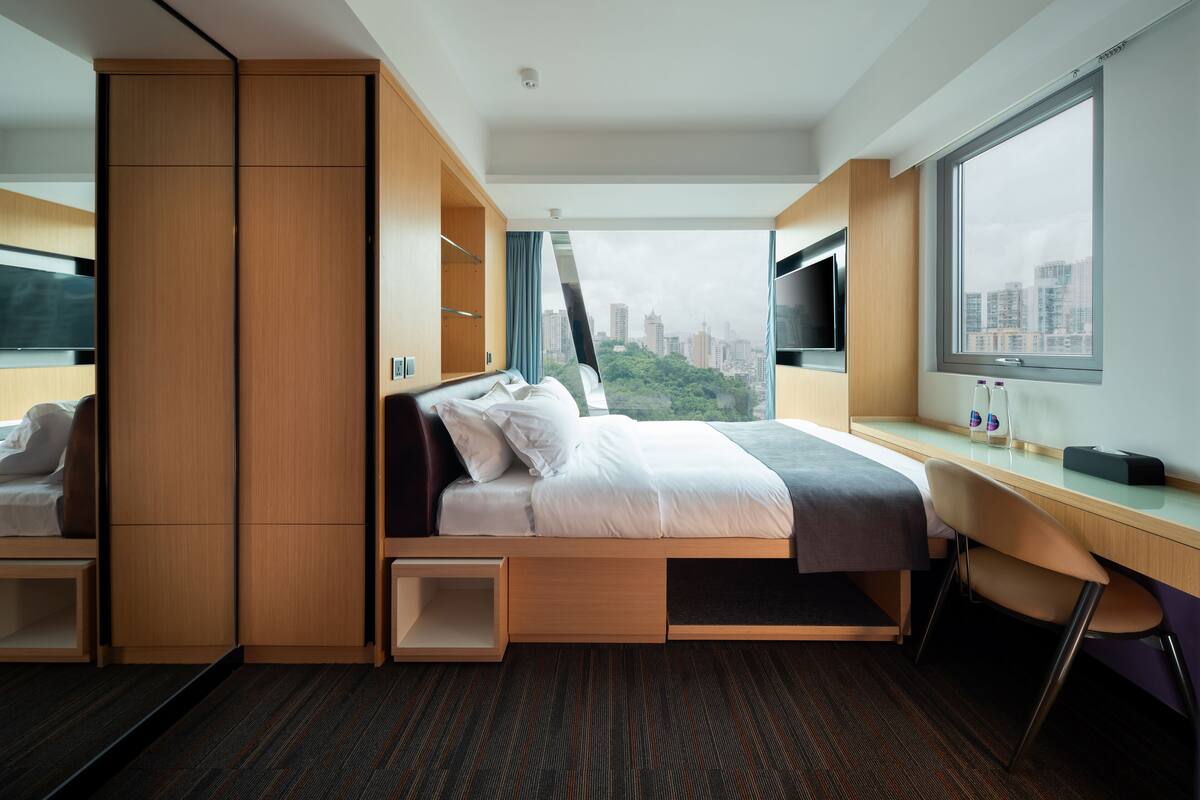 A modern hotel room with a bed, built-in wardrobe, desk, and a large window offering a city view.