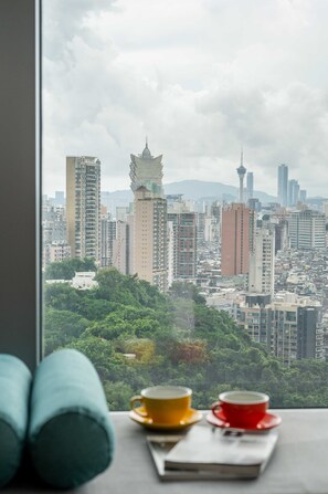A view from a window with two cups on a tray, overlooking a cityscape with tall buildings and greenery. - Greenery Inn (Macau)