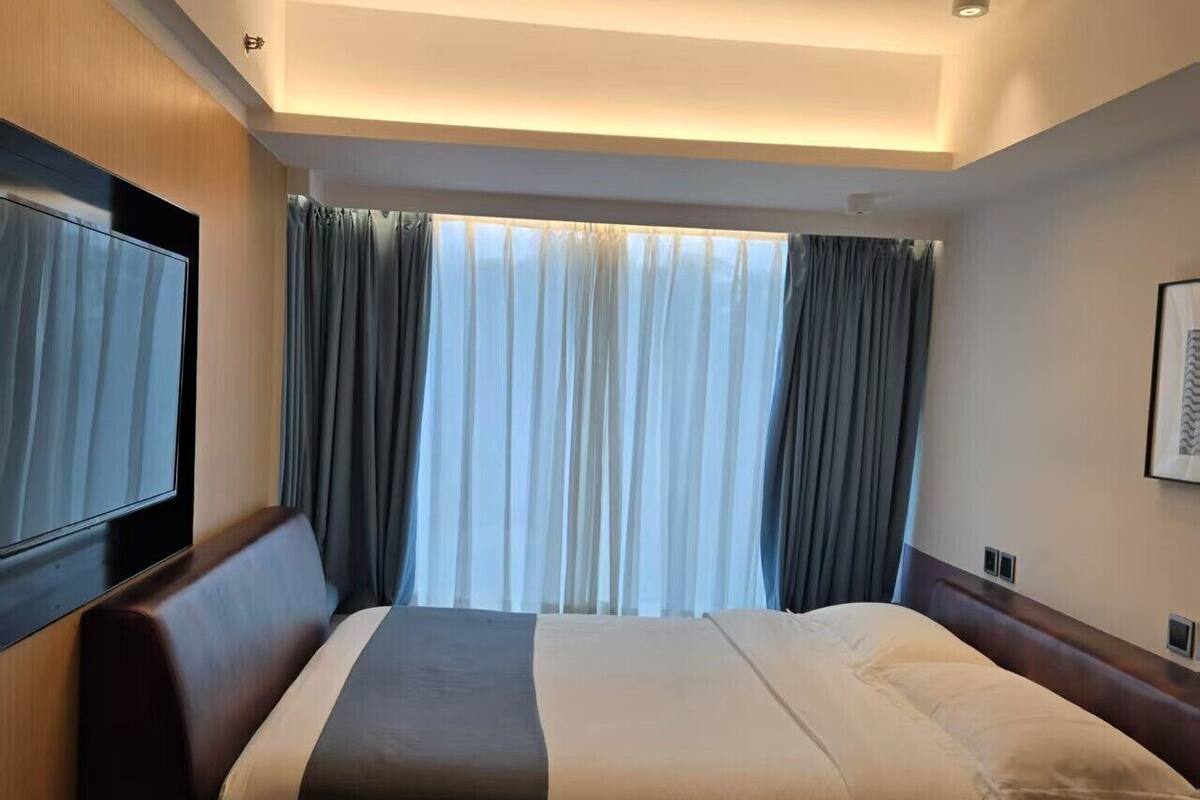 A hotel room with a bed, a TV mounted on the wall, a window with curtains, and a telephone on the side table.