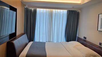 A hotel room with a bed, a TV mounted on the wall, a window with curtains, and a telephone on the side table.