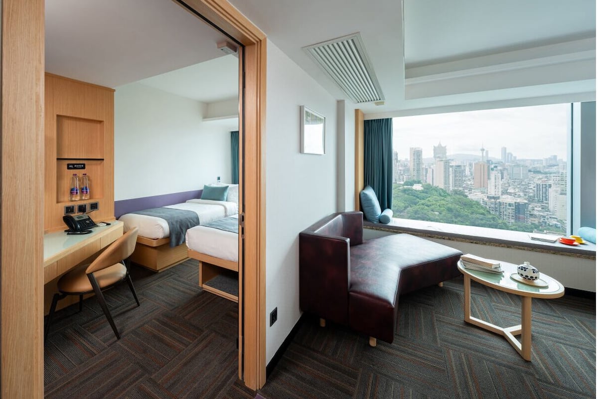 A hotel room with a bed, desk, chair, and a view of the city.