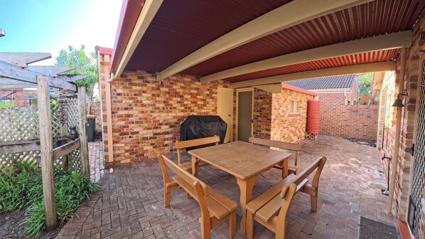 A covered patio with a brick wall, a wooden table with benches, and a barbecue grill.