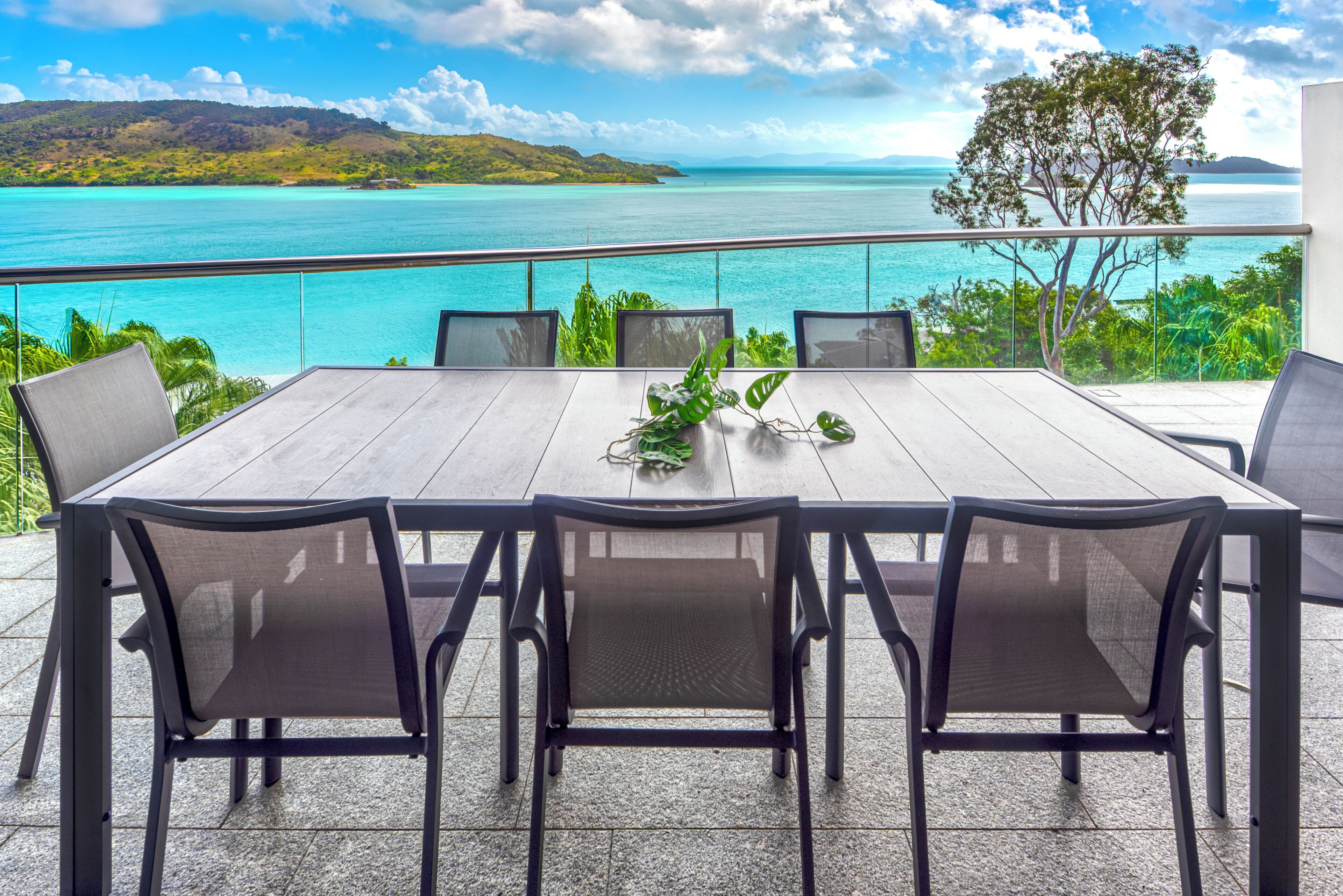 Une table à manger avec six chaises, une balustrade en verre et une vue sur une mer turquoise et une terre lointaine.