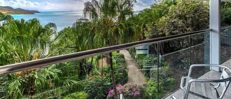 A balcony with a glass railing overlooking a lush garden and a coastal view.