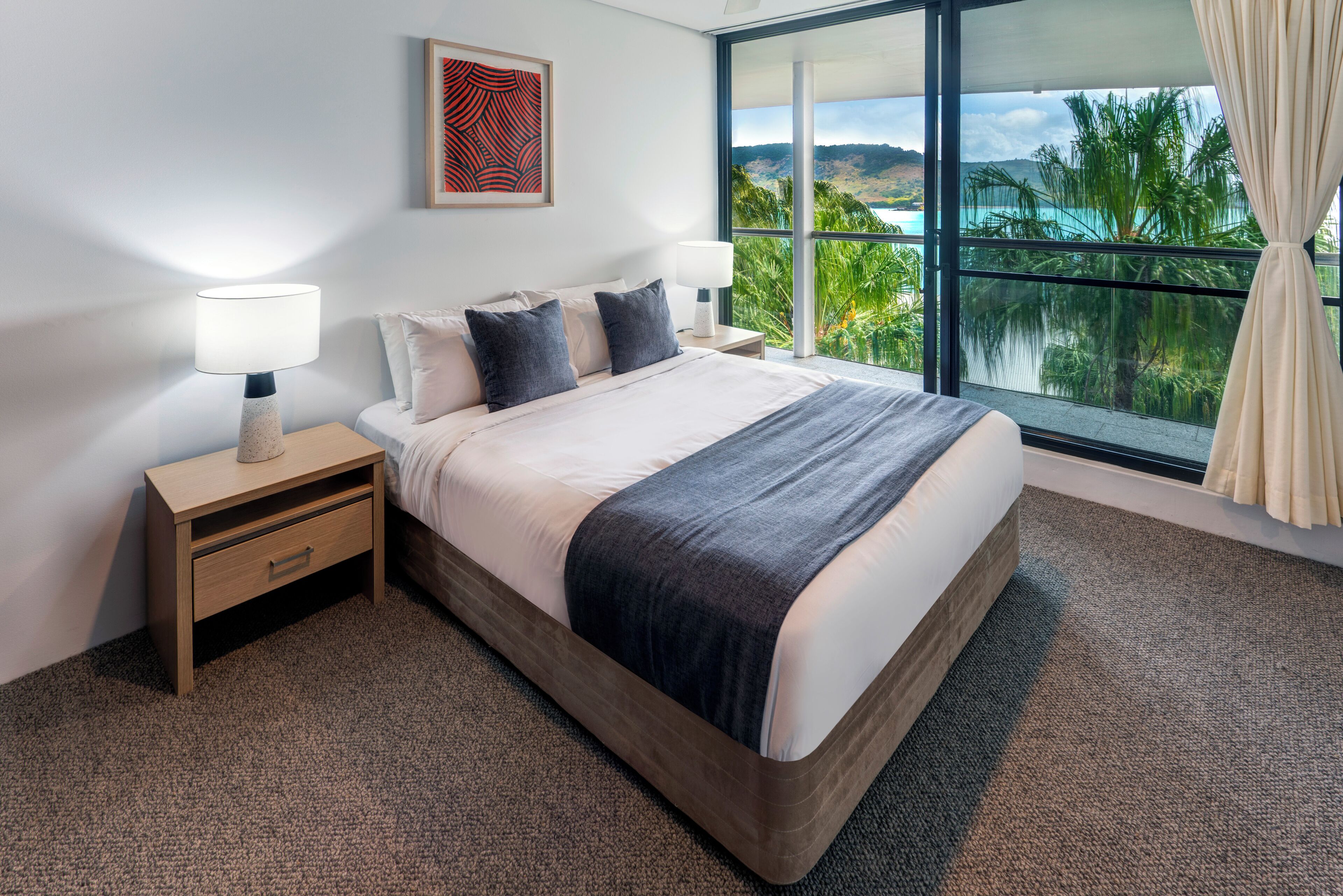A hotel room with a large bed, two bedside tables, a nightstand, a window with a view of the ocean and palm trees, and a framed artwork on the wall.