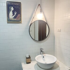 A bathroom with a white tiled wall, a round mirror, a white sink, and a framed poster on the wall.