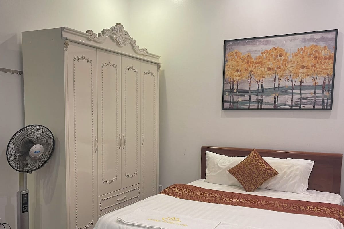 A bedroom with a bed, a wardrobe, a fan, and a painting on the wall.