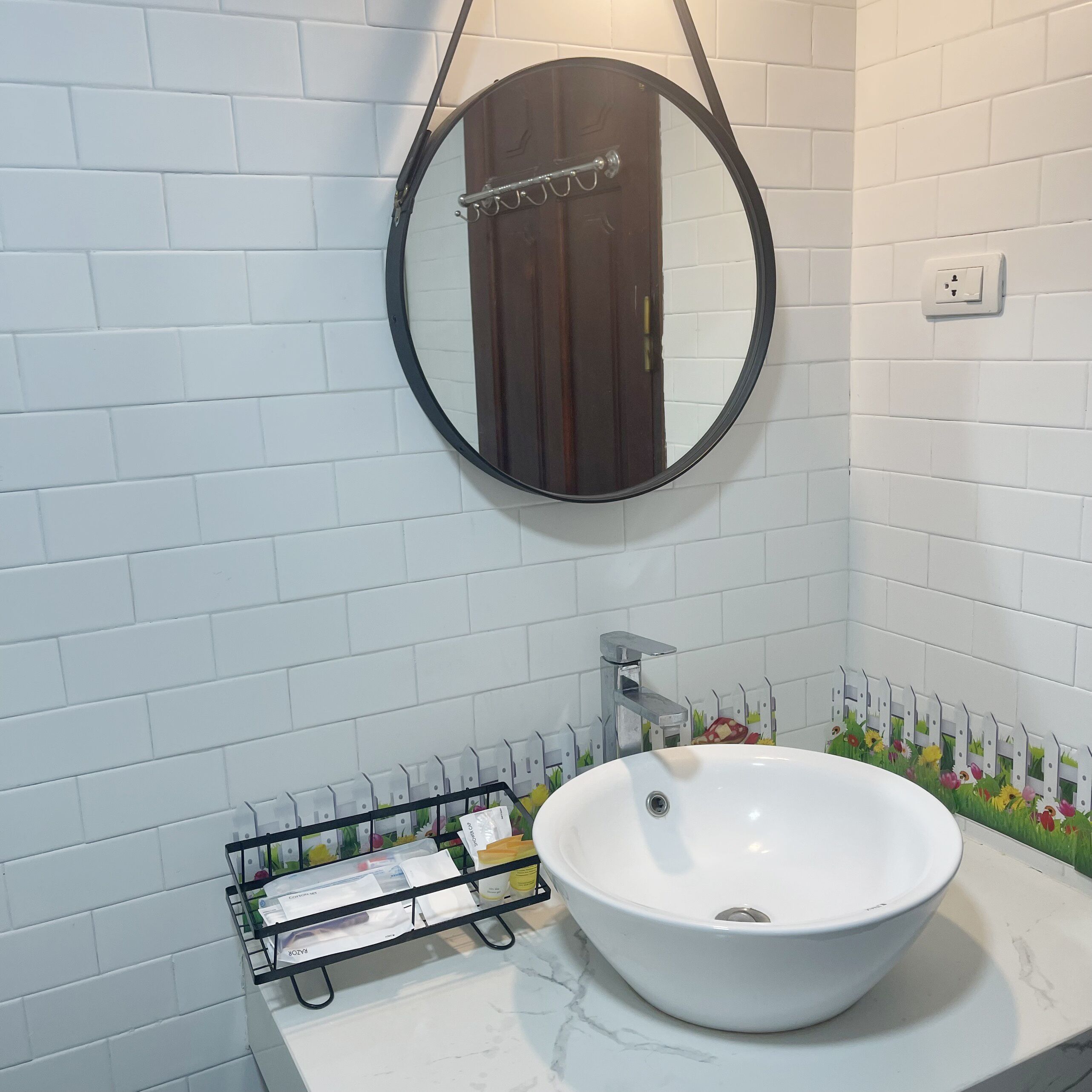Un baño con pared de azulejos blancos, un espejo redondo, un lavamanos blanco con un solo grifo y un perchero con algunas toallas.
