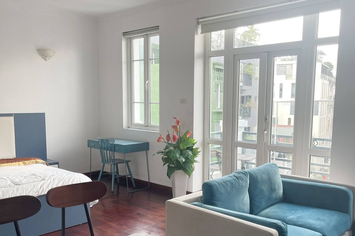 A modern room with a bed, a dining table, a blue sofa, and large windows offering a view of buildings.