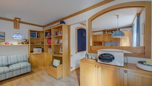 A cozy living room with a sofa, wooden shelves with books, a microwave, and a small kitchen area. - Appartamento Alpetta 37 Ski In - Ski Out (Sestriere)