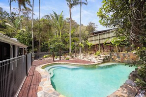 Una piscina rodeada por un solárium de piedra y una valla metálica, con vegetación tropical y un muro de piedra al fondo.