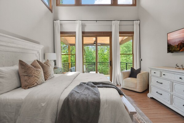 Quarto com cama grande, uma cadeira, cômoda e vista para o verde através de janelas amplas.