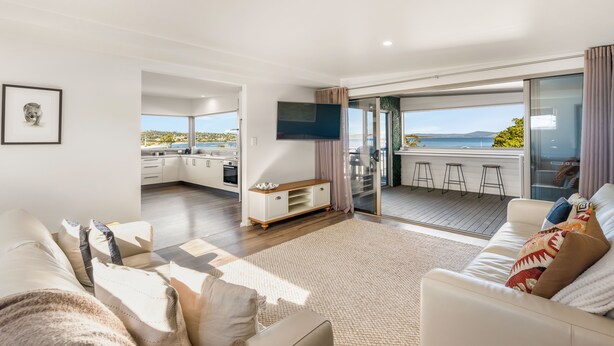 A modern living room with a sofa, a coffee table, and a TV. There is a sliding glass door leading to a balcony with a view of the sea.