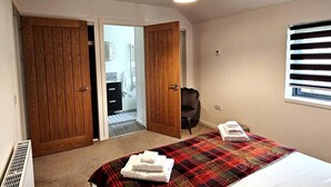 A bedroom with a bed, a red plaid blanket, two towels, a wooden wardrobe, a bathroom with a mirror, and a window with blinds. - Luxury 3-bed House in the Scottish Highlands (Inverness)