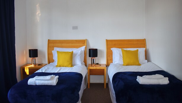 Habitación de hotel con dos camas, cada una con cabecero de madera, ropa de cama blanca y almohada amarilla. Hay dos mesitas de noche con lámparas y toallas dobladas sobre las camas.