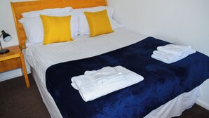 Una cama bien hecha con una colcha azul y dos almohadas blancas, un cabecero de madera y una mesita de noche con una lámpara.
