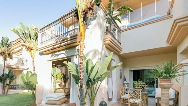 Une maison à deux étages dotée d’un balcon, d’un espace extérieur aménagé pour s’asseoir et de plantes tropicales.