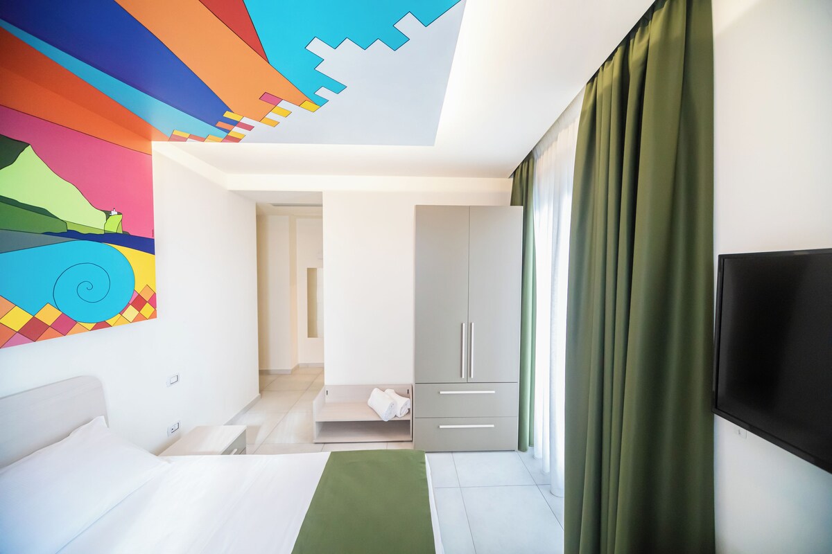Una habitación de hotel moderna con cama, televisor, armario y un cuadro abstracto colorido en la pared.