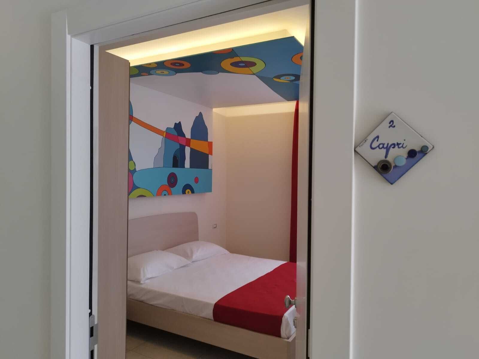 A small, single-bed room with a colorful abstract painting on the wall and a small sign with the name "Capri".