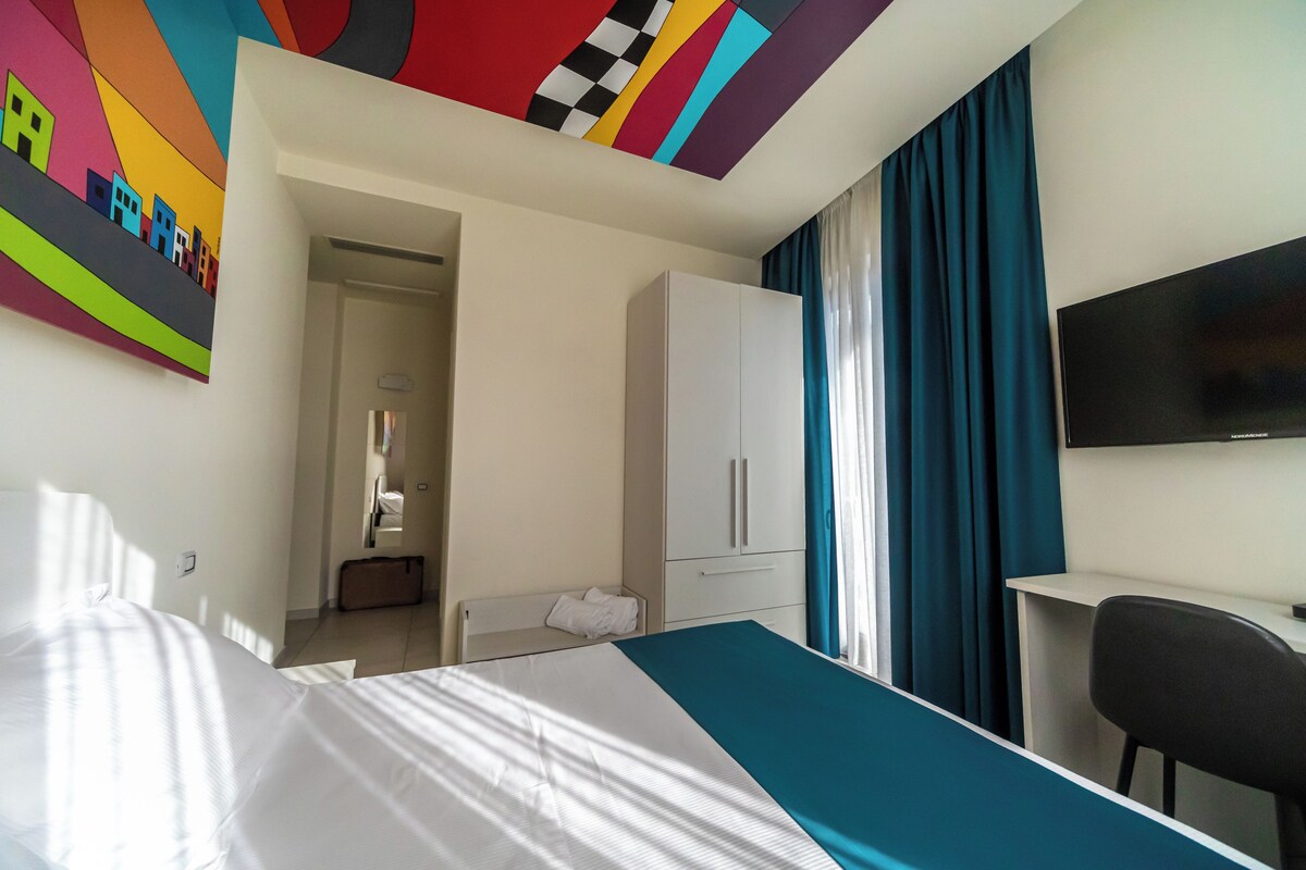 A hotel room with a bed, a desk, a TV, a wardrobe, and a colorful wall art piece.