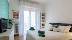 Un dormitorio ordenado con cama, mesita de noche, televisor, estantería y ventana con cortinas.