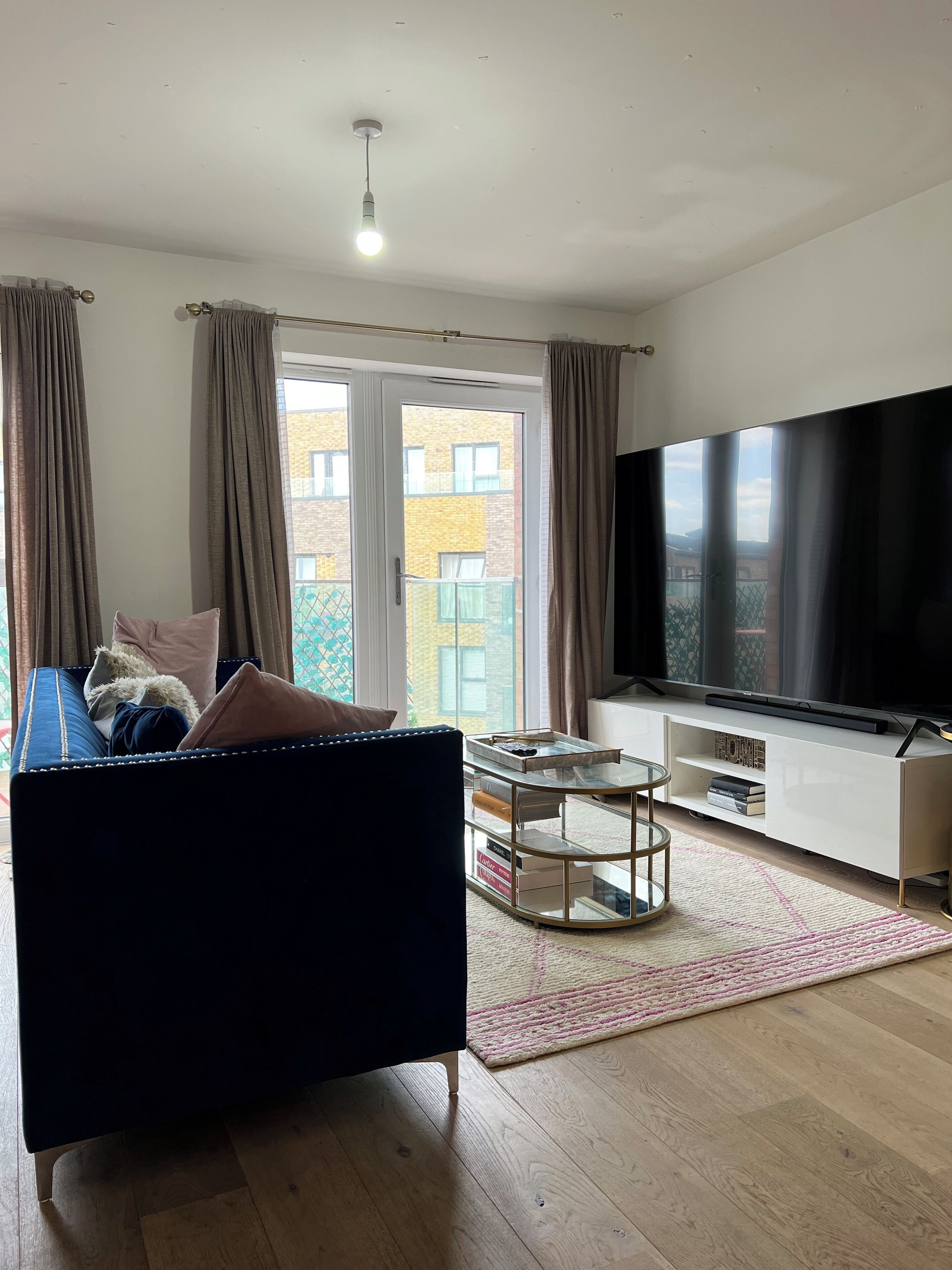 Ein modernes Wohnzimmer mit einem blauen Sofa, einem Glastisch, einem Flachbildfernseher und Blick auf ein Gebäude durch große Fenster.