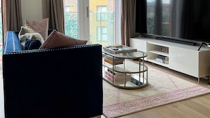 A modern living room with a blue sofa, a glass coffee table, a flat-screen TV, and a view of a building through large windows. - Stunning 1-bed Apartment in Dartford (Dartford)